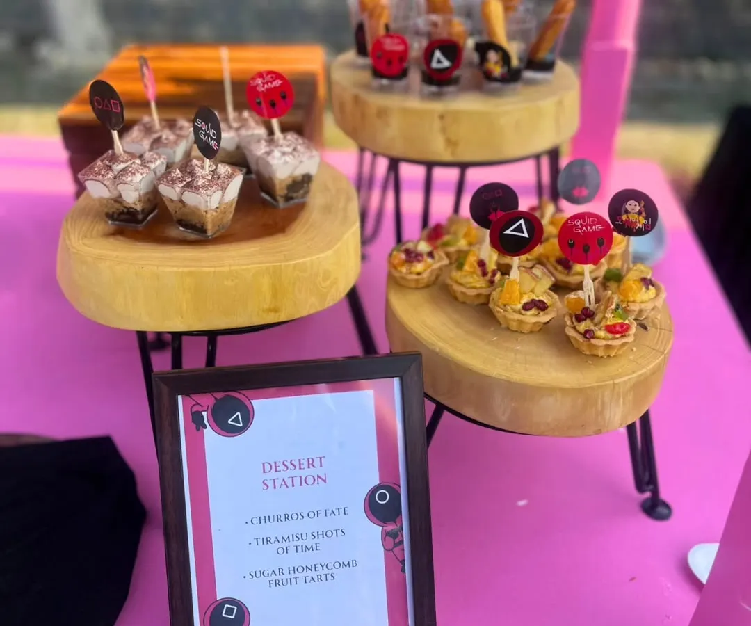 A close-up of the 'Squid Game' themed dessert station, featuring 'Churros of Fate' and 'Tiramisu Shots of Time'. The custom menu and themed toppers show how every detail is designed to fit your party's story.