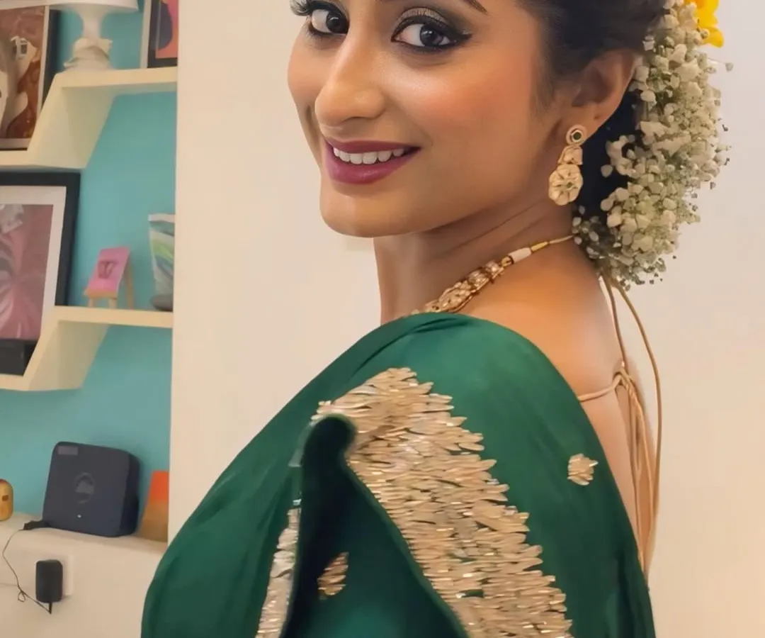 This is a classic engagement look, featuring luminous skin, defined smokey eyes, and a warm lip tone. The hairstyle is an elegant floral bun with soft tendrils framing the face, perfectly complementing the traditional green saree.