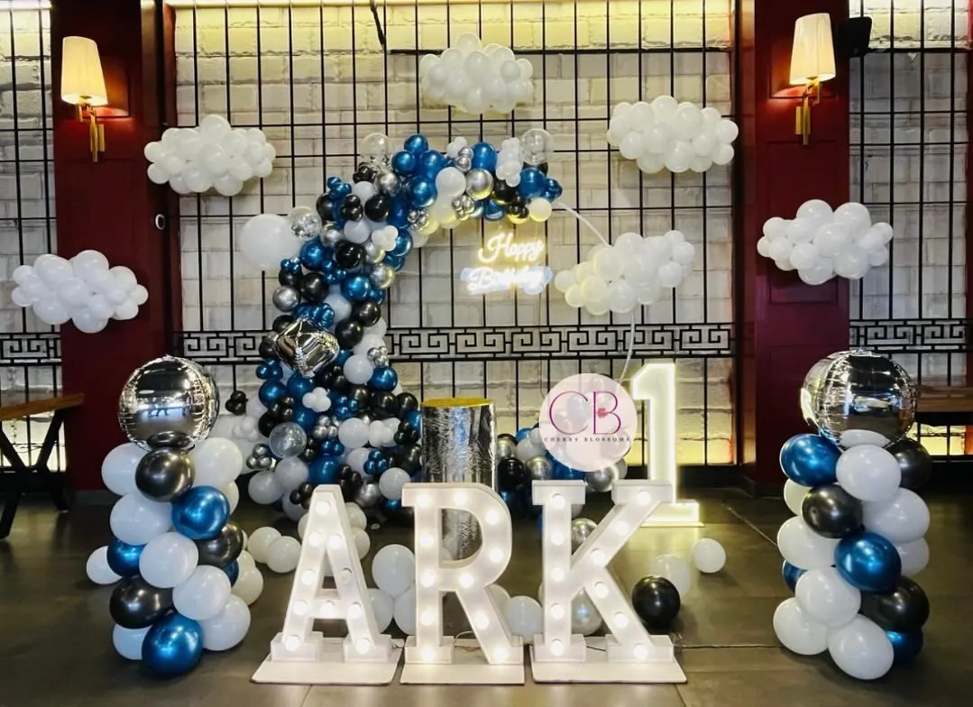 For a classy and simple first birthday, we designed this cloud-themed decor. The setup includes a balloon arch in shades of blue, black, and silver, marquee letters spelling the name 'ARK', and soft cloud-like balloon clusters to create a dreamy atmosphere.