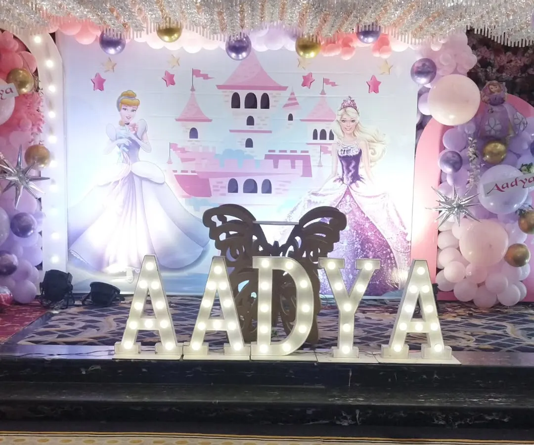 For a magical princess party, I designed this beautiful stage featuring a castle backdrop and two Disney princesses. The large, illuminated marquee letters for the name 'AADYA' create a perfect centerpiece for photos, complemented by soft pink and purple balloon decorations.