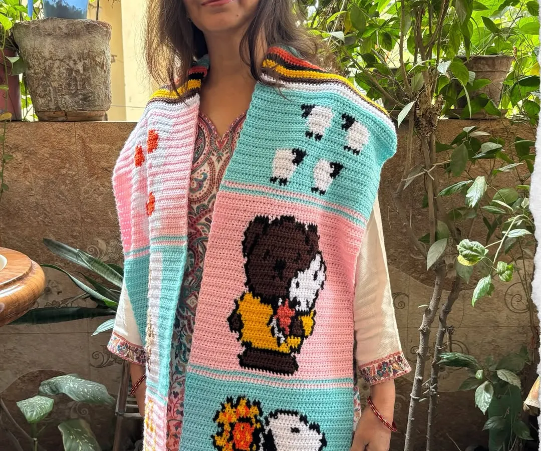 Here is my mom, Nirmala, proudly showing off a one-of-a-kind tapestry muffler we made for a customer. It features fun cartoon characters like Snoopy, sheep, and ducks, all hand-stitched with love. We can create a custom story just for you.