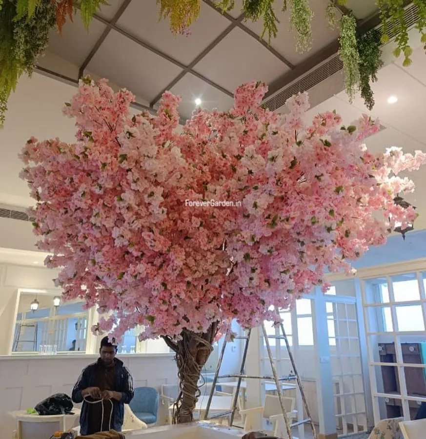 This stunning, large-scale artificial pink cherry blossom tree acts as a magnificent centerpiece in a cafe, offering a vibrant and unique ambiance that attracts customers.