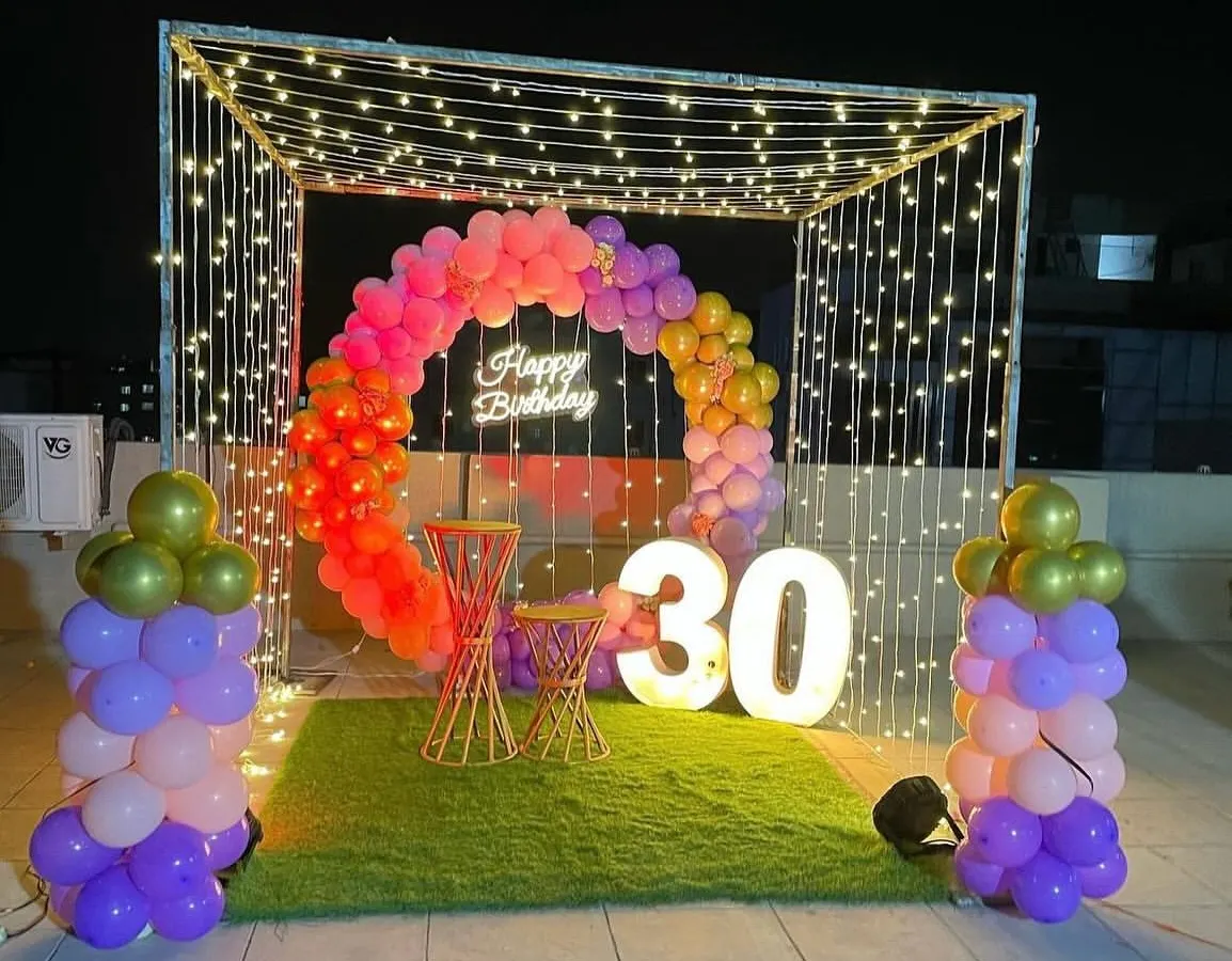 I transform terraces into stunning party spaces, like this 30th birthday celebration. The decor includes a canopy of fairy lights, a colorful balloon ring, a "Happy Birthday" neon sign, and large light-up marquee numbers.