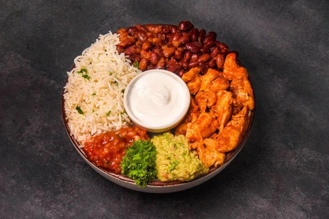 A vibrant and healthy chicken burrito bowl, neatly arranged with cilantro lime rice, seasoned chicken, kidney beans, fresh guacamole, and salsa. The top-down flatlay composition on a dark surface highlights each fresh ingredient.