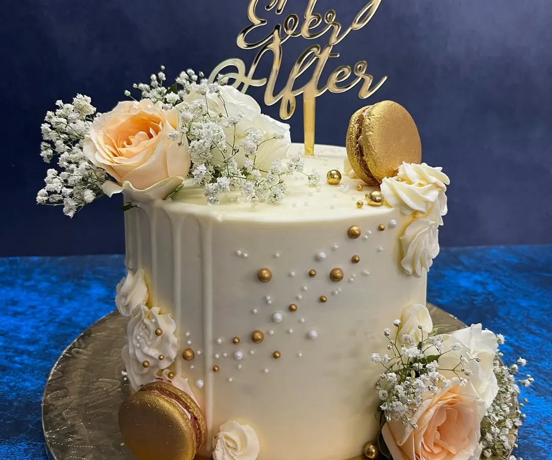 A classic white drip cake for a 'Happily Ever After' moment. This elegant design is adorned with fresh peach roses, baby's breath, and shimmering gold macarons, perfect for engagements, anniversaries, or intimate weddings.