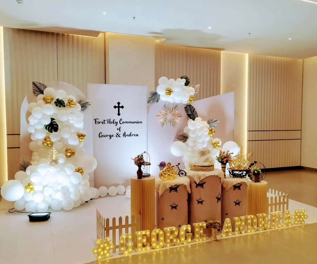 For George and Andrea's First Holy Communion, we designed a sophisticated and modern stage. The decor combines clean white panels, elegant balloon clouds, and touches of gold, with personalized marquee lights to make the day truly special and blessed.