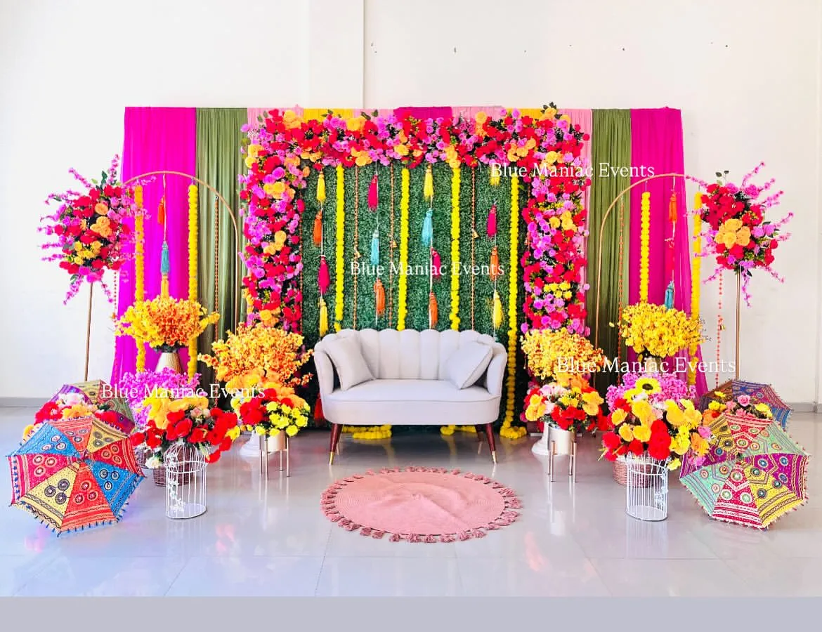 For a vibrant Mehendi or Haldi, I can transform any space with a burst of color. This grand setup uses a lush green wall as a base, framed by a riot of pink, yellow, and red artificial flowers, with traditional umbrellas and floral arrangements completing the festive look.