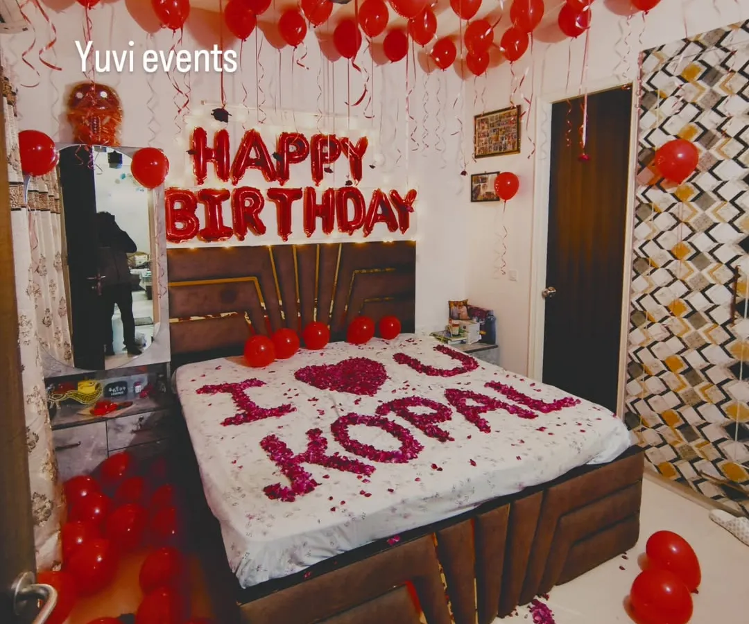 This is my classic romantic birthday room decoration. The setup includes red helium balloons covering the ceiling, 'HAPPY BIRTHDAY' foil letters above the bed, and a personal message like 'I ❤️ U' written with fresh rose petals on the bed.