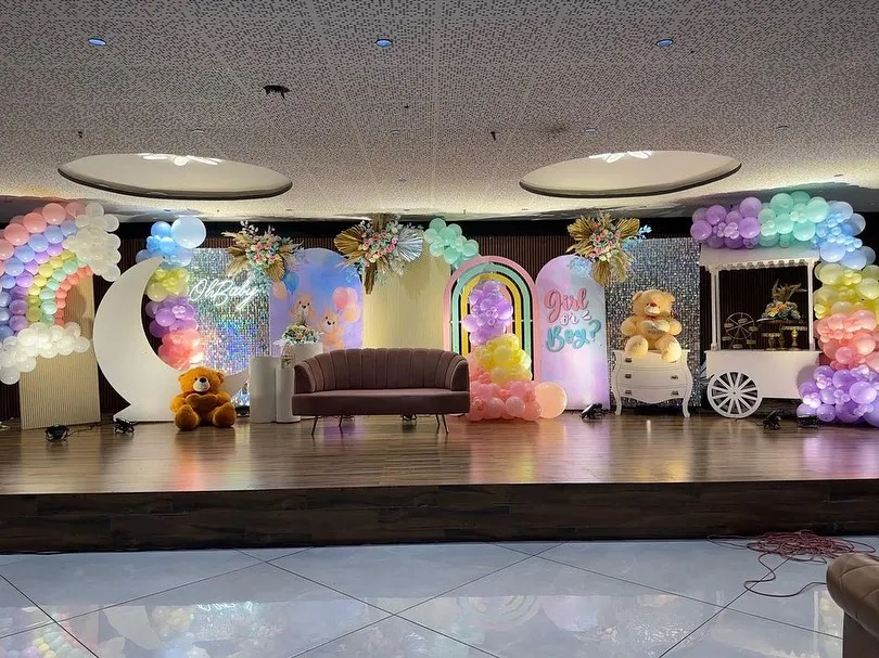 A full view of a spectacular pastel rainbow stage I designed for a large baby shower. The setup features a "Girl or Boy?" reveal sign, a crescent moon prop, multiple backdrops, and a decorative candy cart, all tied together with a vibrant color scheme.