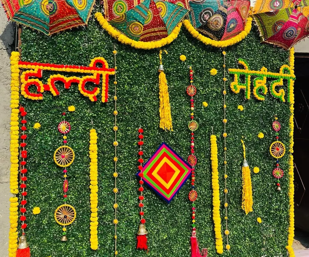For a modern take on traditional Haldi and Mehndi decor, I use an artificial green wall as a base. This backdrop is decorated with marigold garlands, colorful tassels, and Rajasthani umbrellas to create a vibrant and festive photo corner.
