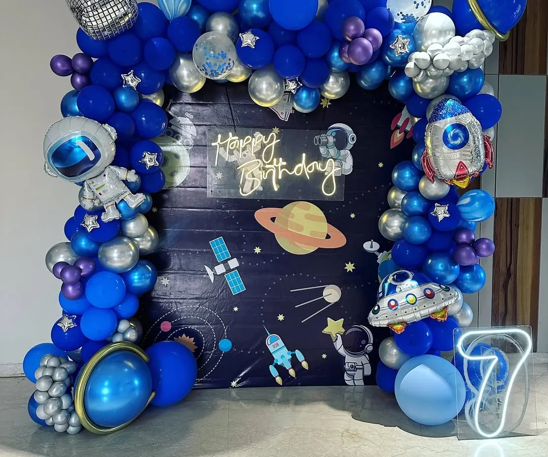 An out-of-this-world space theme birthday party for a 7-year-old. This detailed setup includes a galaxy backdrop, a full balloon arch in shades of blue and silver, and fun foil balloons shaped like astronauts, rockets, and planets.
