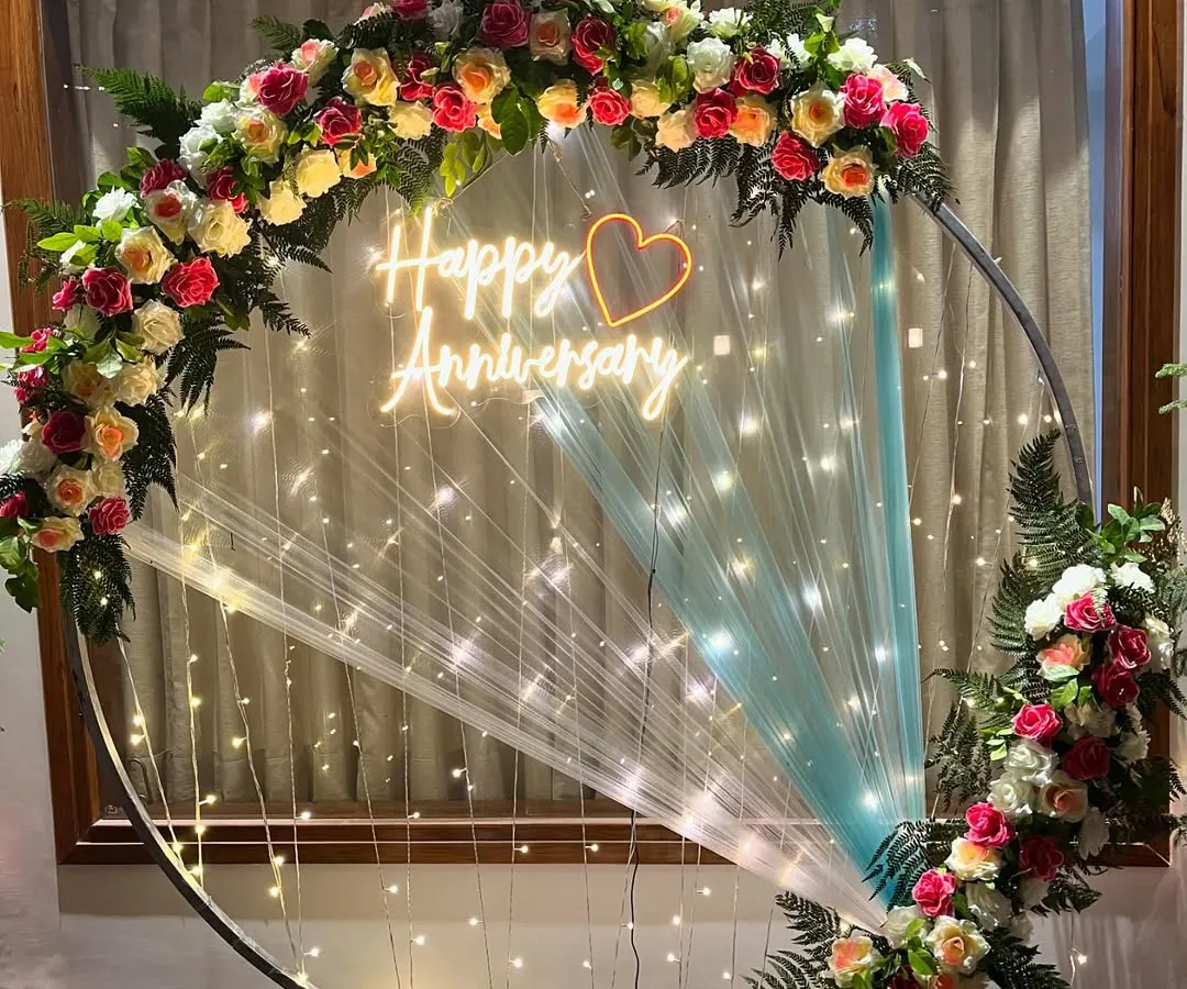 An elegant ring backdrop perfect for an anniversary. This setup features a metal ring decorated with fresh, colorful flowers, delicate fairy lights, and a custom 'Happy Anniversary' neon sign, creating a beautiful and romantic focal point.