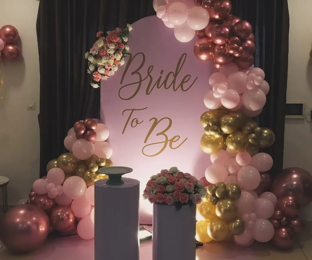 This is a chic bridal shower decoration I created using a popular pink chiara panel backdrop with custom gold 'Bride To Be' lettering. The look is completed with an organic balloon garland in shades of pink, gold, and maroon, and plinths for the cake and floral arrangements.