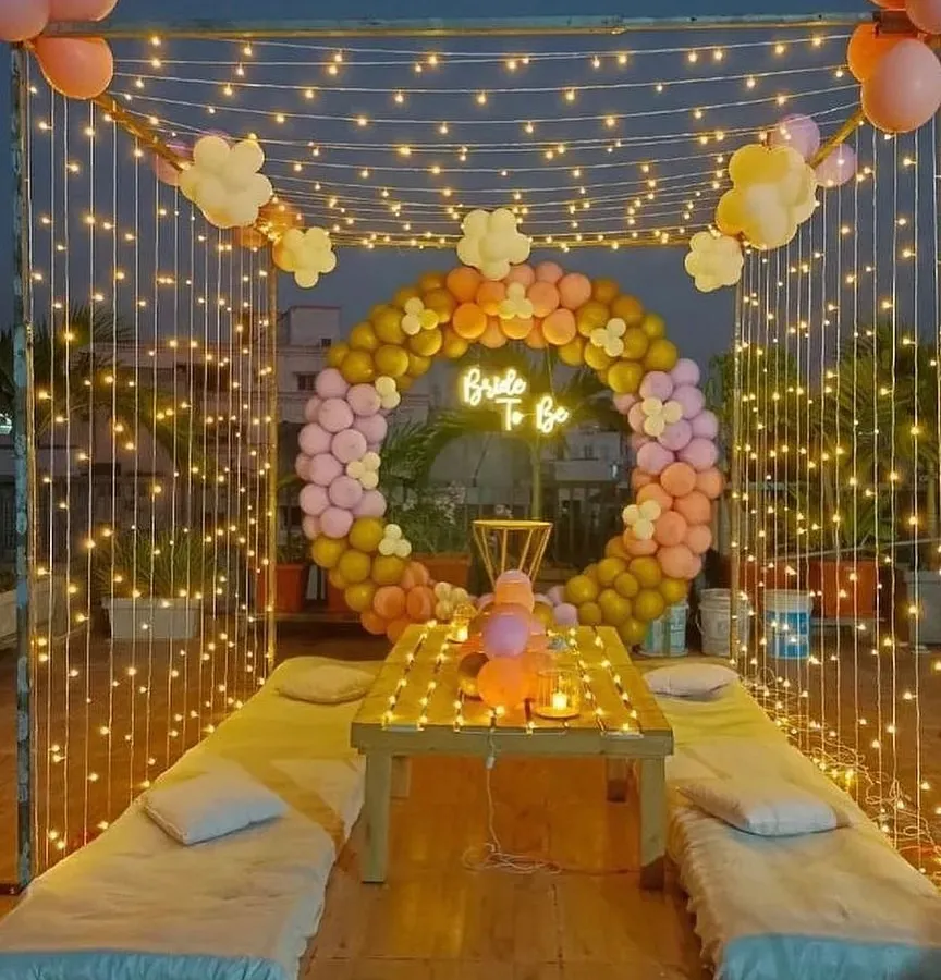 I created this magical bride-to-be party on a terrace, perfect for an evening celebration. The setup includes a canopy of fairy lights, low seating with cushions, and a circular balloon arch in pastel shades. A 'Bride to Be' neon sign serves as the centerpiece for photos.