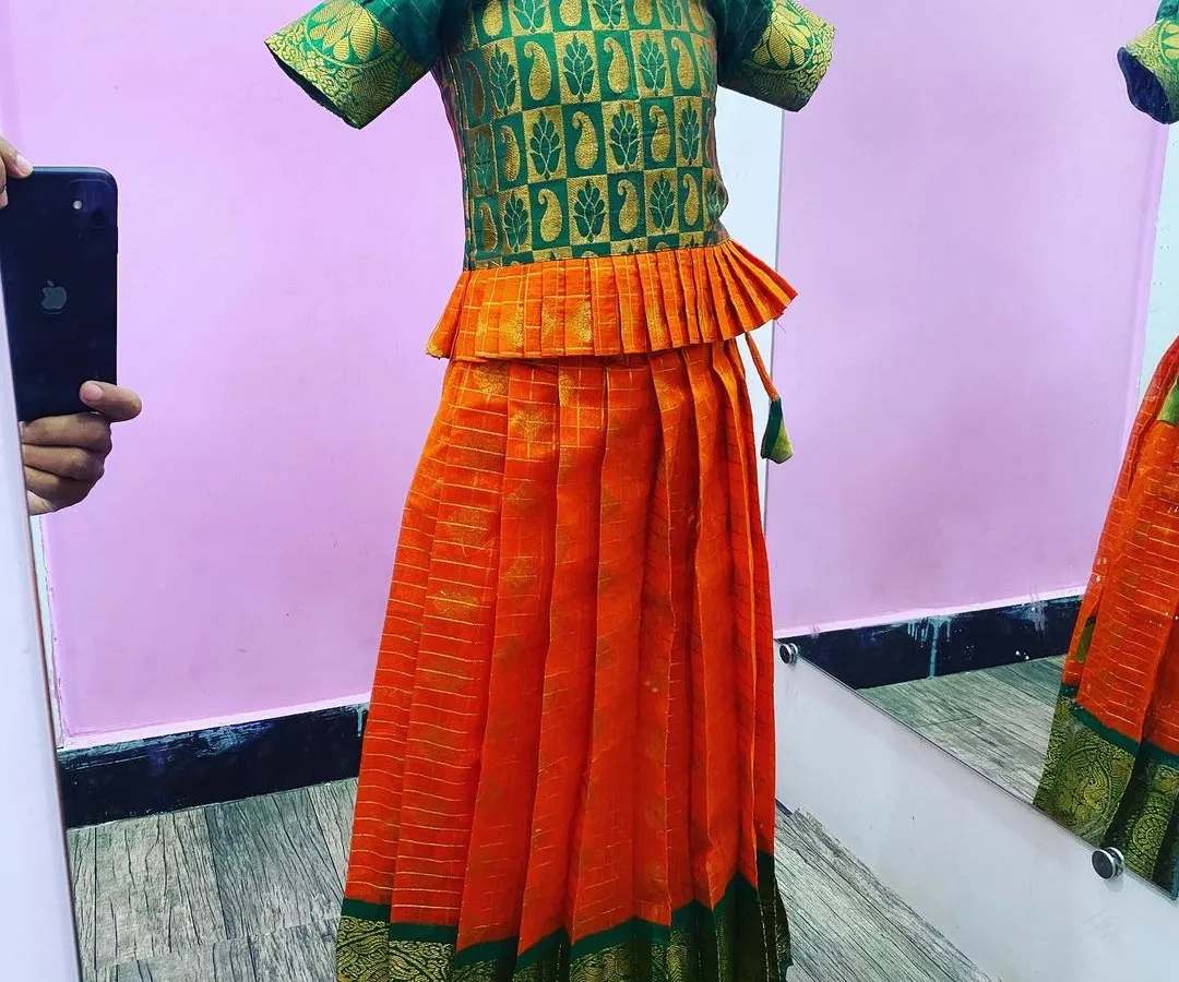 This is a classic kids' Paavada blouse set in a bright orange and green silk combination. The top has a peplum style and puff sleeves, all made with very good stitching for a clean look.