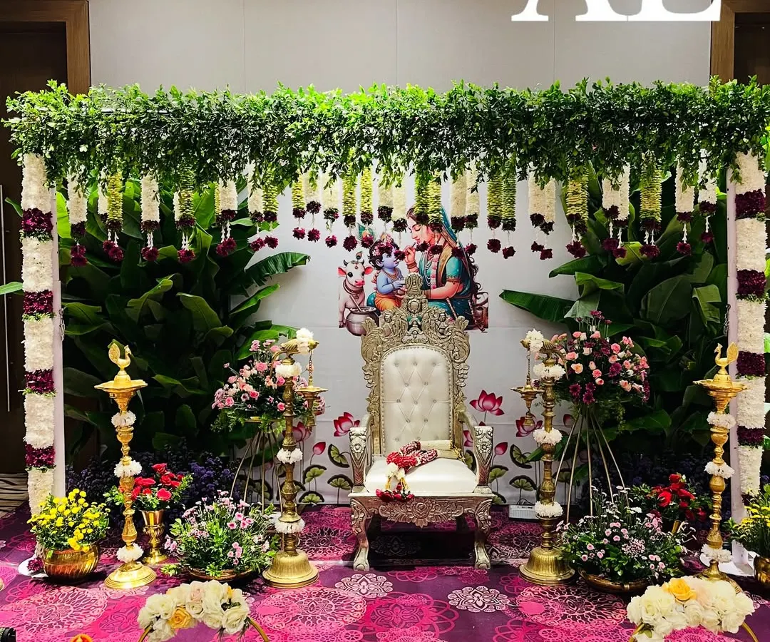 A complete Seemantham (baby shower) setup. This traditional arrangement features a backdrop of fresh banana leaves and jasmine strings, a decorated chair for the mother-to-be, and brass lamps, all surrounded by floral offerings.