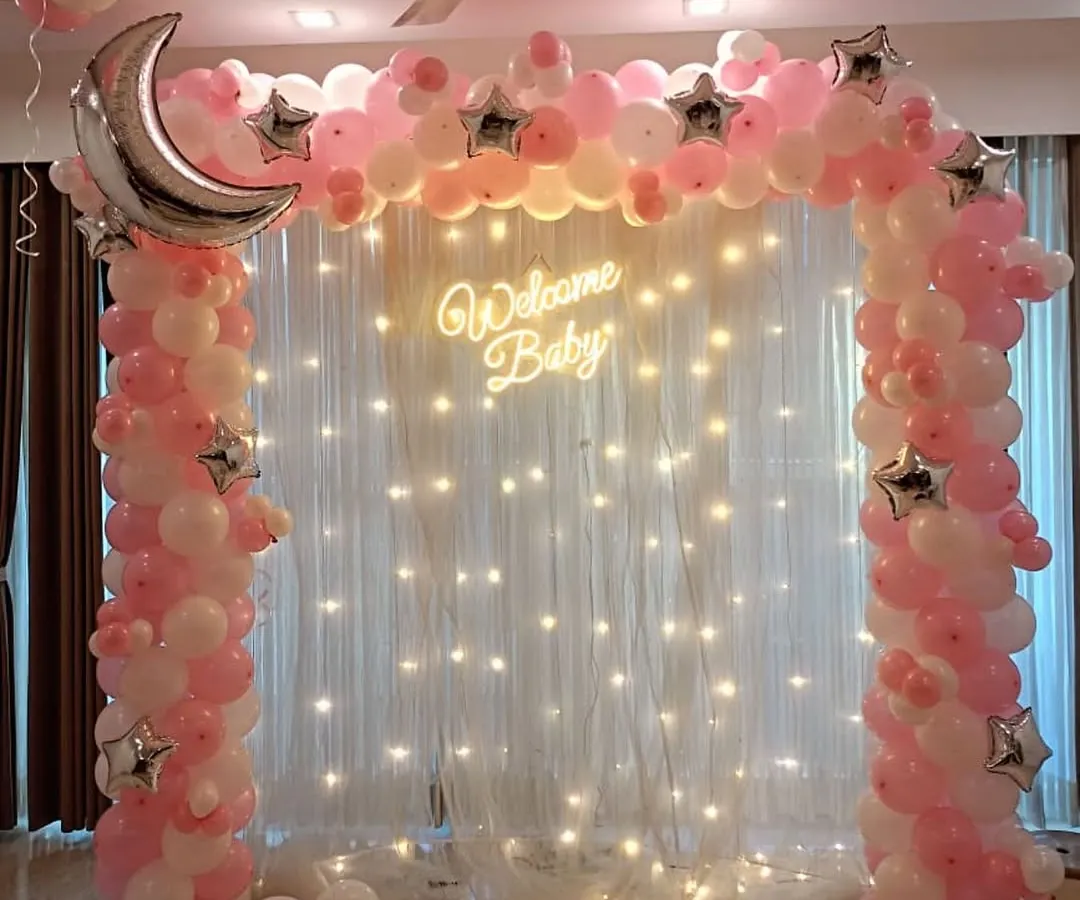 A dreamy 'Welcome Baby' setup perfect for bringing your little girl home. We created this magical moon and stars theme using a pink and white balloon arch, a large silver crescent moon foil balloon, and a backdrop of twinkling fairy lights for a truly special celebration.