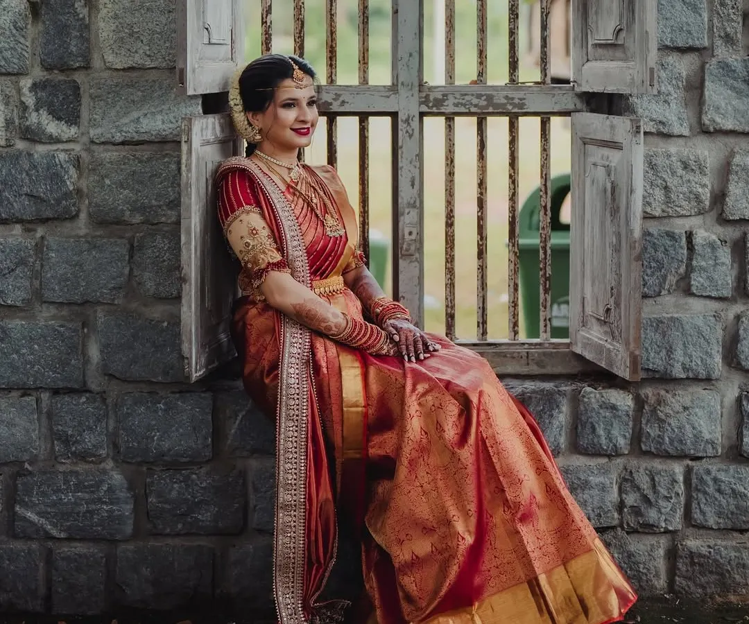 A moment of quiet elegance. This bride is the picture of grace in a traditional red and gold Kanjeevaram silk saree, paired with a bespoke embroidered blouse. The rich texture of the silk and the detailed craftsmanship of the blouse create a timeless bridal look.