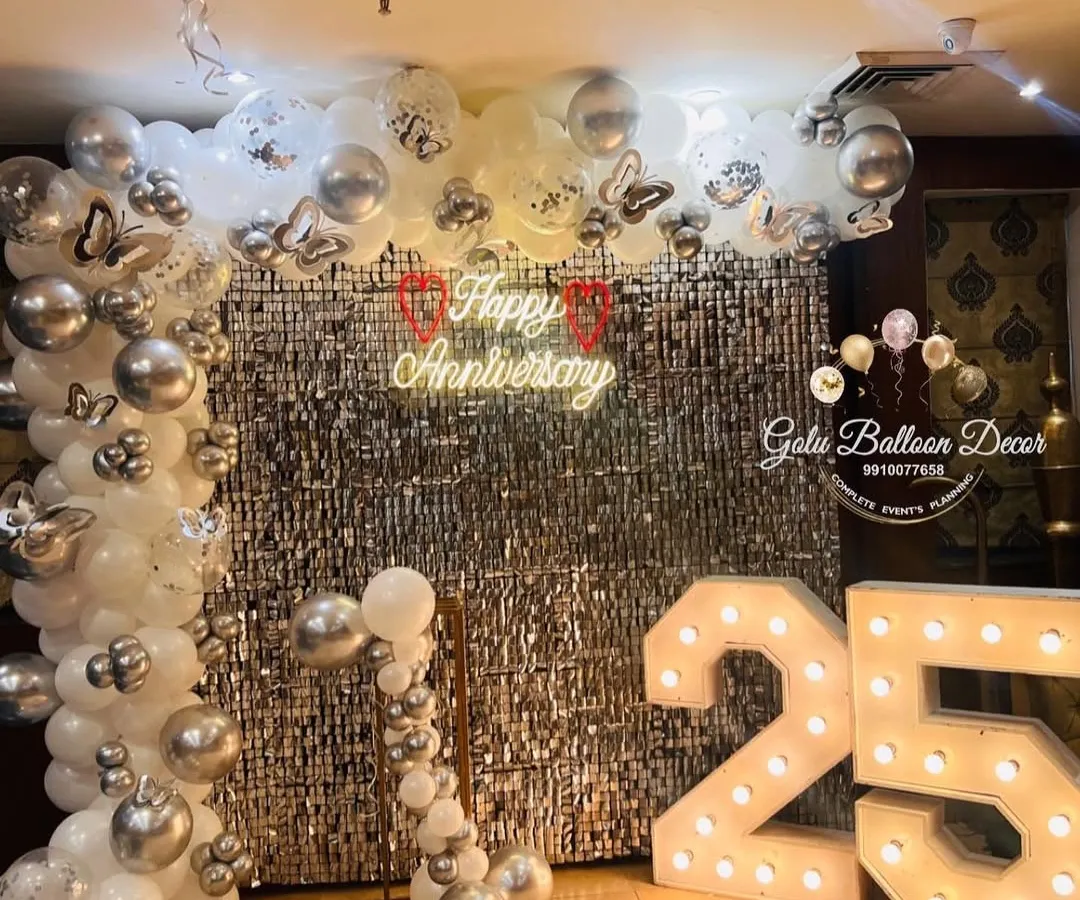 Celebrating a silver jubilee in style. This 25th-anniversary decor features a shimmering silver sequin wall, a custom "Happy Anniversary" neon sign, and large light-up marquee numbers, all framed by an elegant white and silver balloon arrangement.