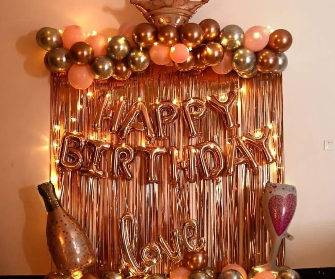 A very popular birthday decoration in a rose gold and gold theme. This setup includes a shiny foil curtain, 'Happy Birthday' letters, a balloon arch topped with a crown, and other fun balloons like a champagne bottle and a 'Love' sign.