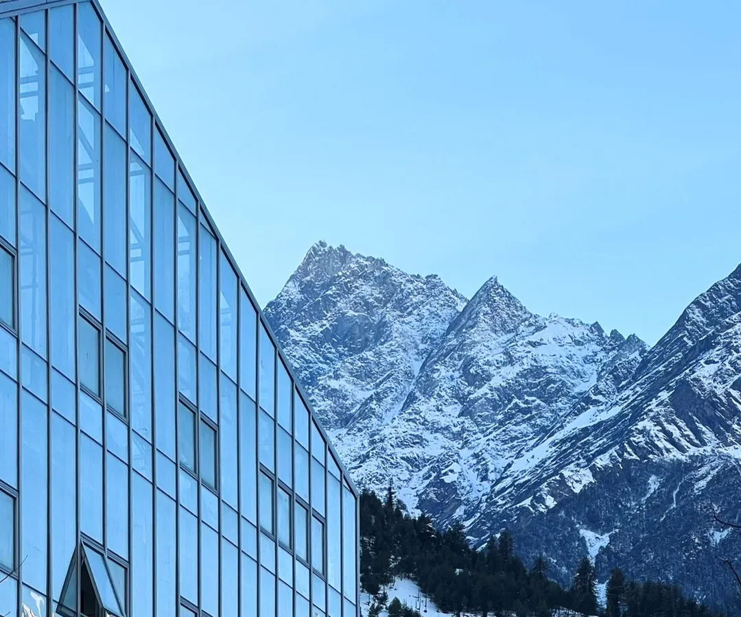 The Hamsa Resort in Manali features a modern glass and steel facade that reflects the surrounding Himalayan peaks. This design choice creates a direct visual dialogue between the structure and its environment, while the building itself incorporates traditional Kathkuni construction principles.