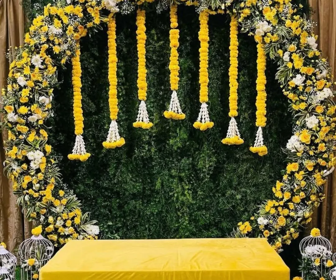 This modern Haldi backdrop features a full floral arch of yellow and white blossoms against a lush green wall. The clean design and comfortable seating create a chic and joyful atmosphere perfect for photos.