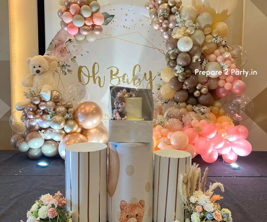 An elegant and modern "Oh Baby" themed baby shower backdrop. The setup features a sophisticated palette of rose gold, pink, and beige balloons, a plush teddy bear, custom pedestals for the cake, and delicate floral arrangements.