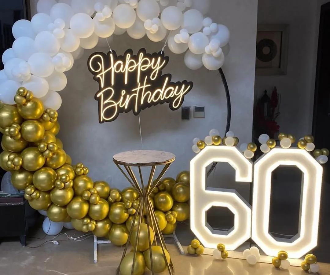An elegant and classic 60th birthday celebration setup. We used a sophisticated gold and white color scheme with a large balloon ring, a modern neon 'Happy Birthday' sign, and prominent light-up marquee numbers to mark the special milestone.