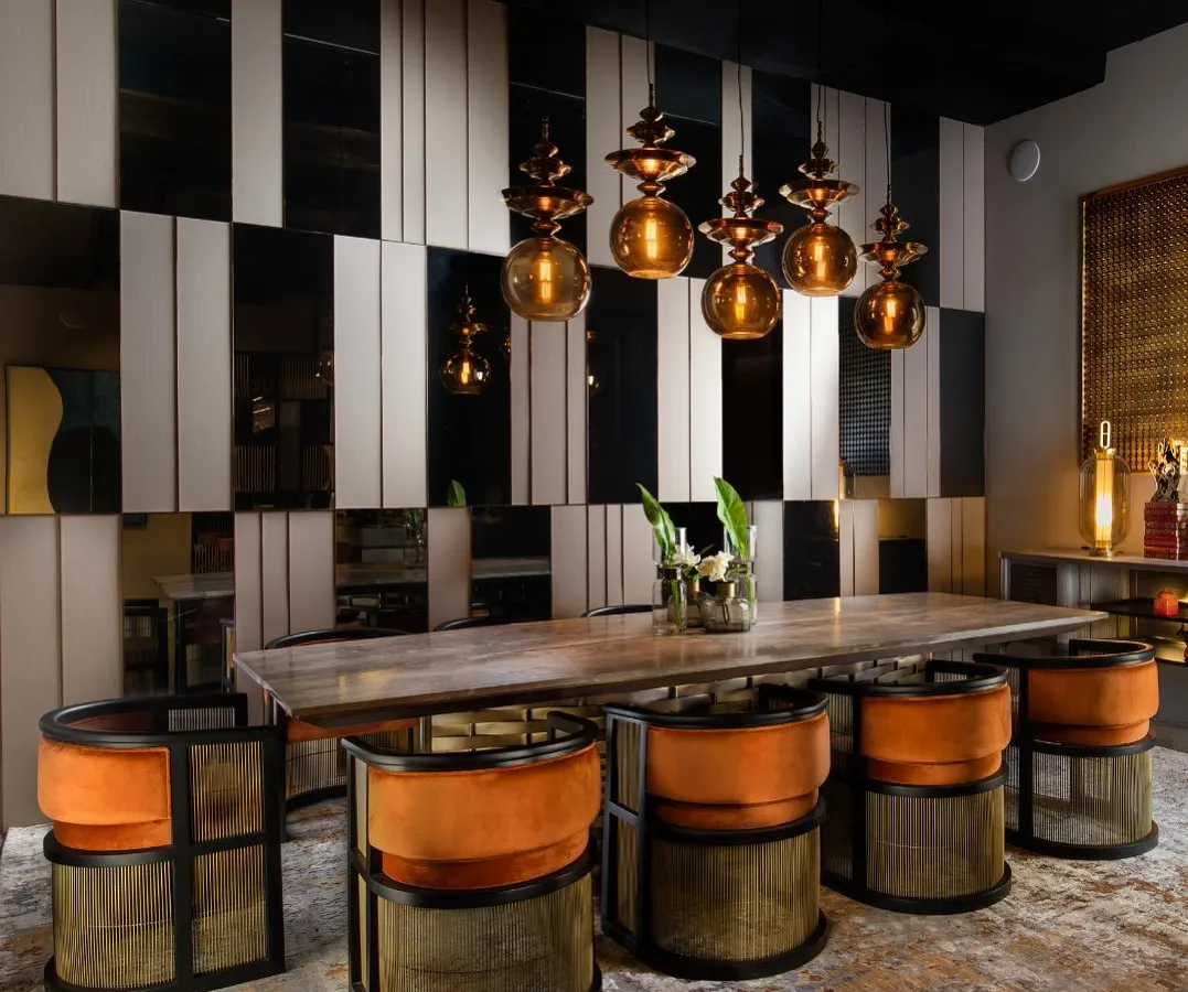 We designed this dining space to be an experience of texture and contrast. The chairs, upholstered in a vibrant tangerine velvet, feature a base of woven metal sheets in a distressed bronze finish, complementing the solid stone tabletop and the dramatic wall paneling.