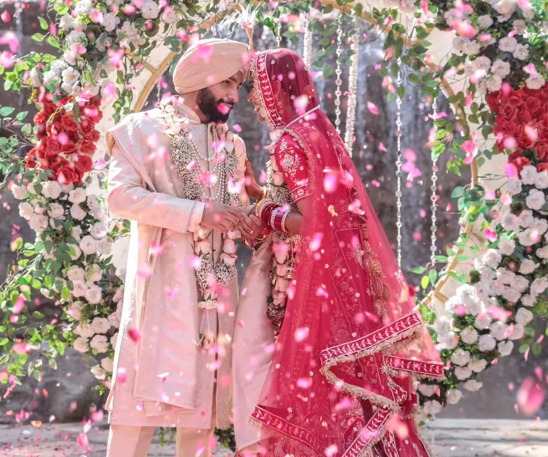 A magical moment as a Sikh couple shares their first look under a stunning floral arch, with a cascade of pink petals showering down around them. This image perfectly captures the joy and emotion of the Varmala ceremony.
