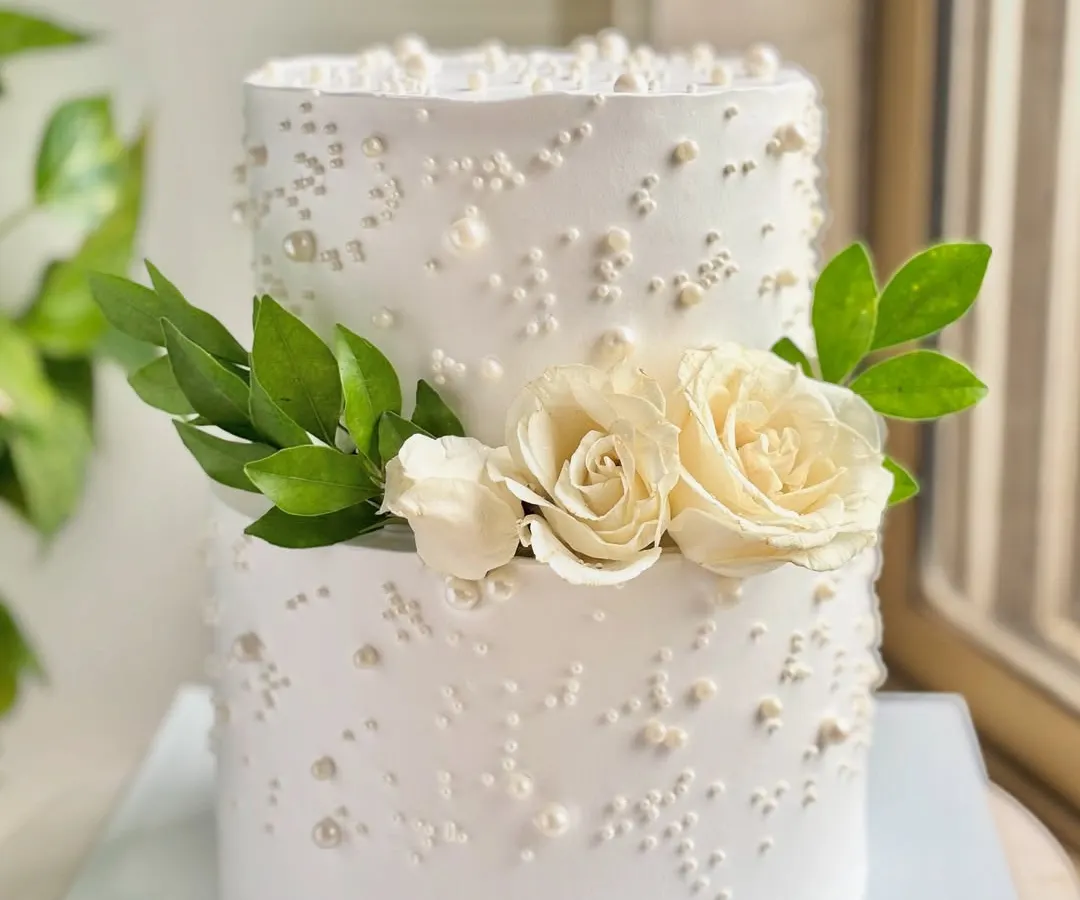 A vision in white, this two-tiered cake is the definition of timeless elegance. The clean buttercream finish is adorned with scattered edible pearls and fresh white roses, making it a perfect centerpiece for a sophisticated roka or wedding celebration.