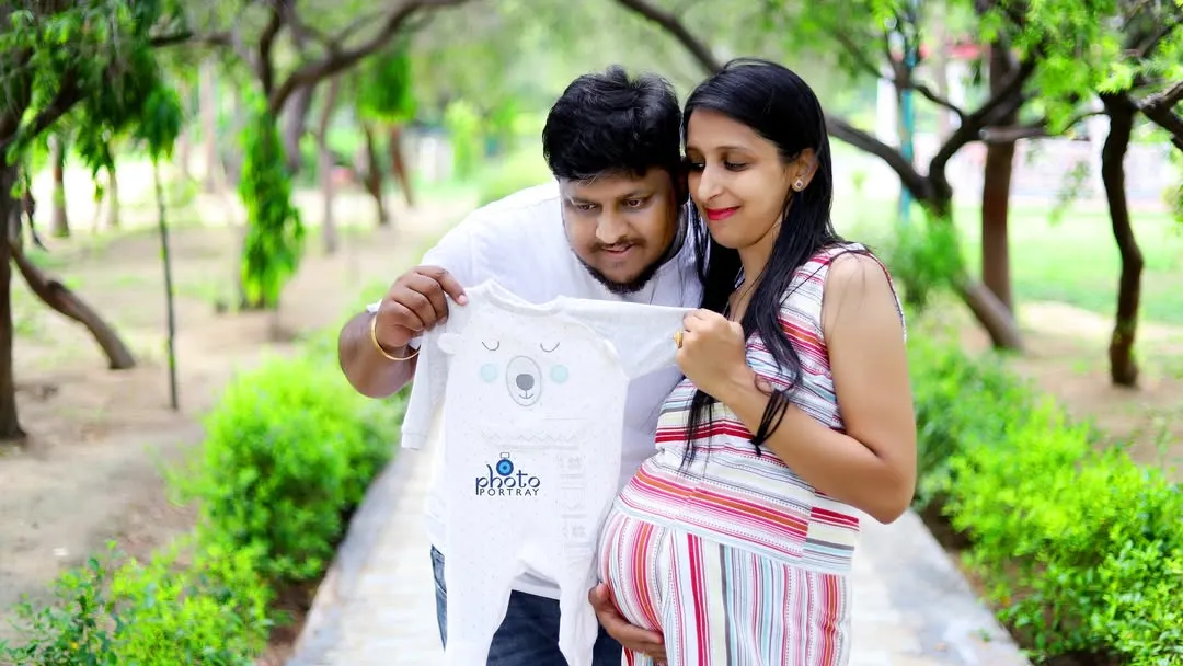 An expecting couple shares a quiet moment in a park, looking at a baby onesie together. This image from an outdoor maternity photoshoot in Noida captures the sweet anticipation of welcoming a new family member.