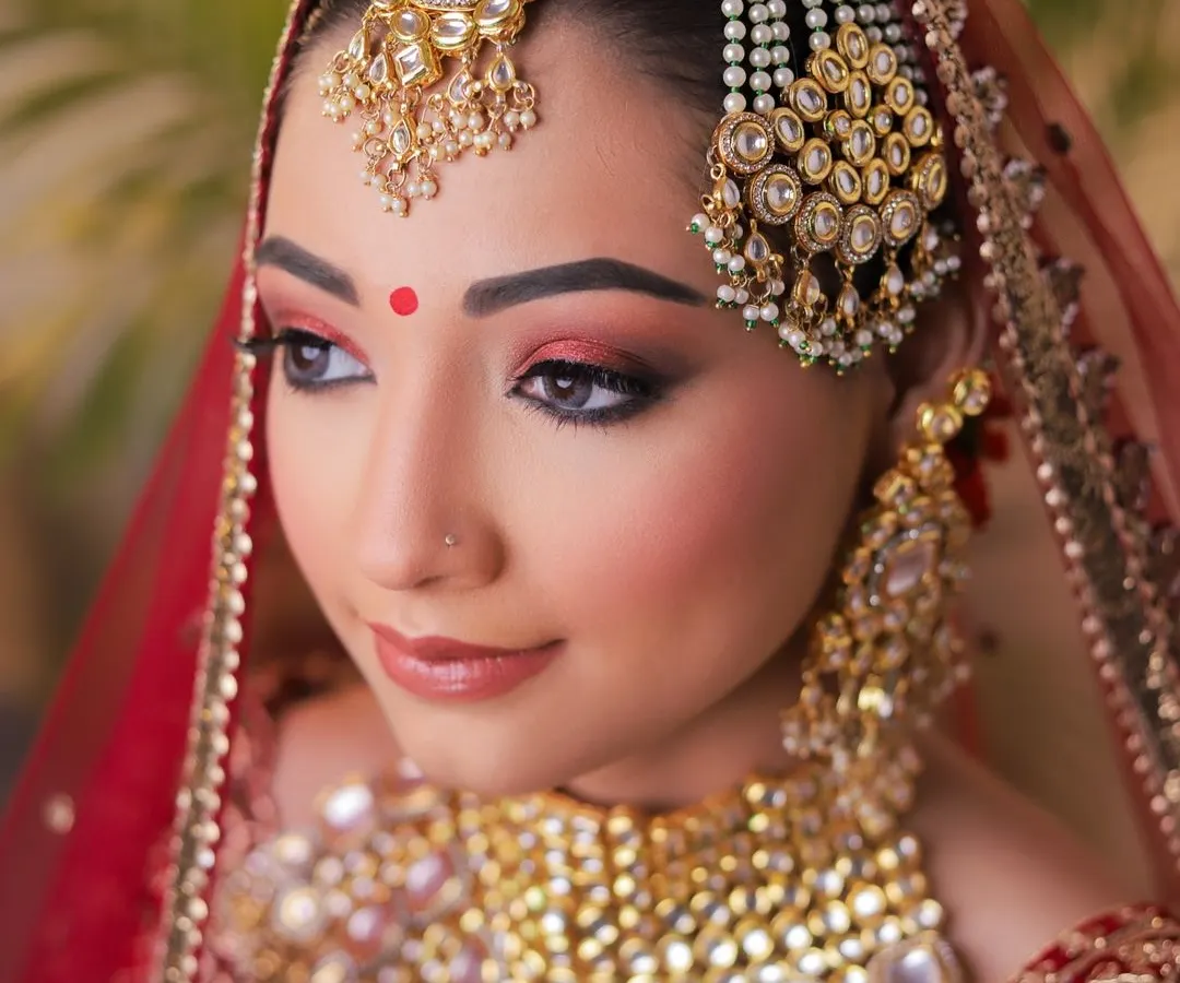 A close-up of a classic Indian bridal makeup look. This shot focuses on the intricate details, from the warm, smoky red eyeshadow and perfectly defined brows to the elaborate kundan and pearl jewellery, including the maang tikka and passa. The soft lighting ensures the makeup looks flawless and vibrant.