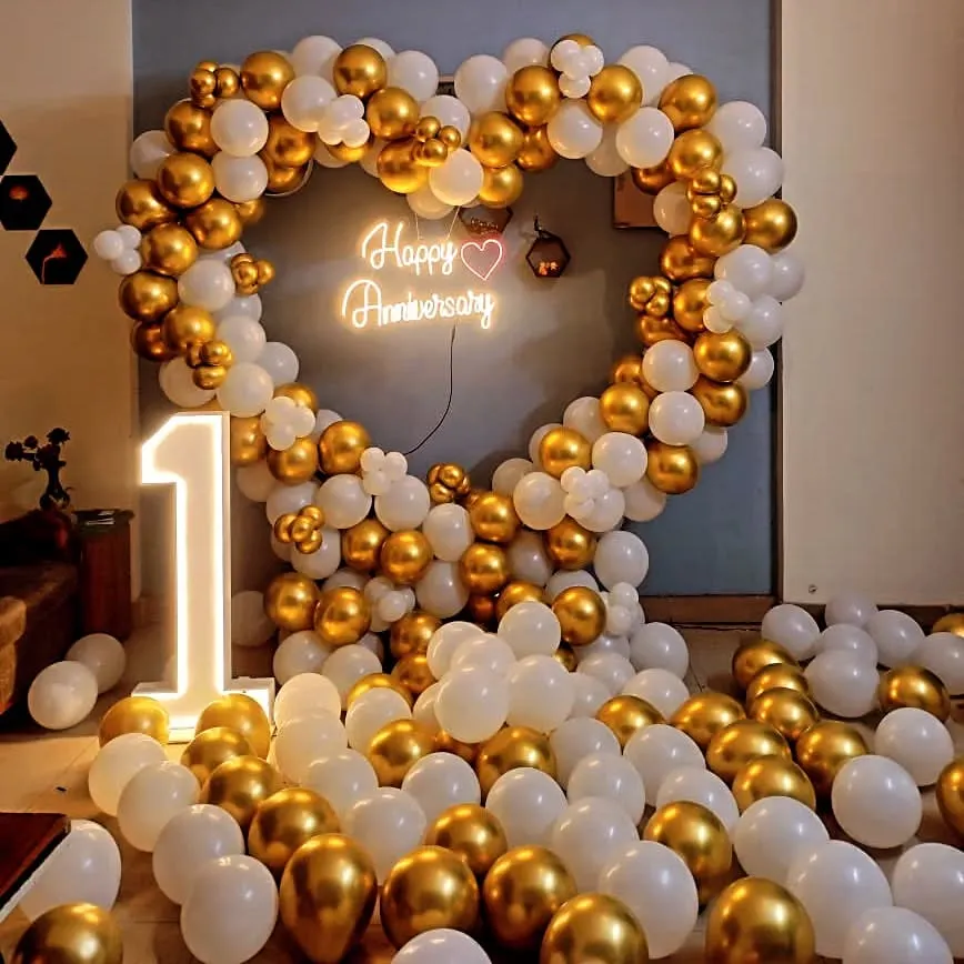 An elegant and romantic first-anniversary decoration. This setup features a large heart-shaped balloon arch in chrome gold and white, a custom 'Happy Anniversary' neon sign, and a light-up number '1', perfect for an intimate and stylish celebration.