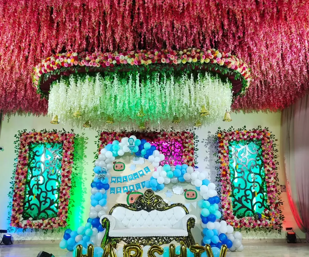 This grand stage was designed for a 'Welcome Baby' ceremony, featuring an ornate white throne-style sofa for the family. The backdrop includes a dense balloon arch in shades of blue and white, custom banners, and large gold foil balloons spelling out the baby's name, all under an elaborate ceiling of hanging flowers.