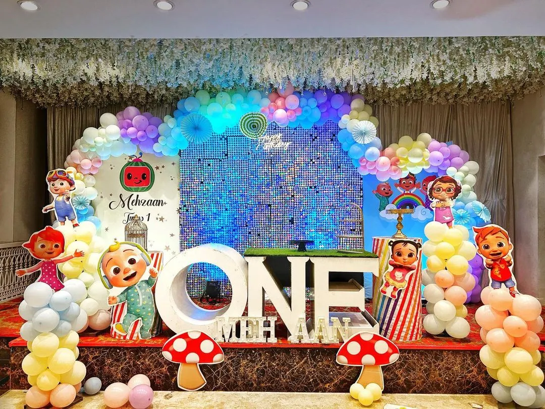 A spectacular Cocomelon themed first birthday party stage. This vibrant setup includes a full cast of character cutouts, a massive "ONE" prop, a shimmering backdrop, and a rainbow of balloon decorations.