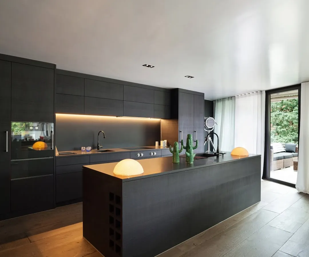 This modern pantry and breakout space was created for a corporate office, featuring sleek, handleless cabinetry and a large central island. Warm, under-cabinet lighting and natural wood floors balance the dark, minimalist aesthetic, providing a welcoming area for employees to connect.