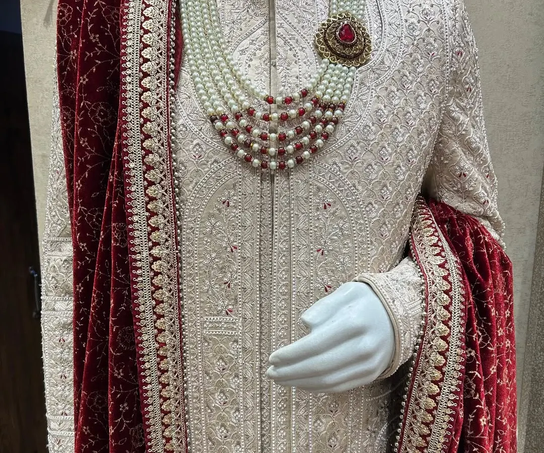 The epitome of royal elegance. This is a close-up of our classic ivory sherwani, paired with a rich red velvet stole and a multi-layered pearl and ruby necklace, showcasing the detailed zardosi and threadwork for a truly grand wedding look.