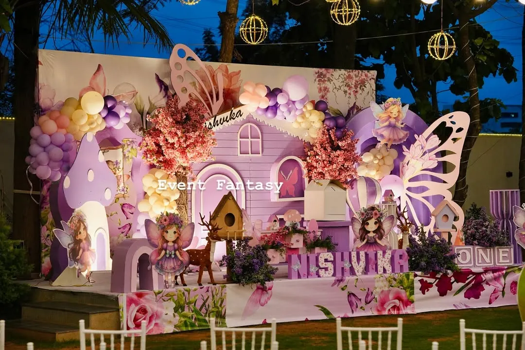 A magical fairy garden party for Nishvika, glowing under the evening sky. This enchanting setup includes a whimsical purple fairy house, large butterfly wing props, custom name lettering, and soft lighting from hanging lanterns to create a dreamlike atmosphere.