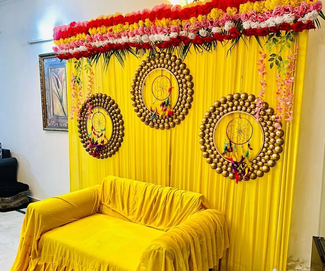 For a home mehndi or haldi function, I can create a vibrant photo corner like this one. This setup includes a bright yellow backdrop with matching sofa covers, decorative dreamcatcher-style hangings, and a floral border on top.