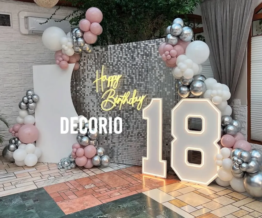 An exciting 18th birthday celebration with a glamorous silver sequin wall that creates a stunning photo opportunity. The setup includes large, illuminated '18' marquee numbers, a 'Happy Birthday' neon sign, and a balloon arch in shades of pink, silver, and white.
