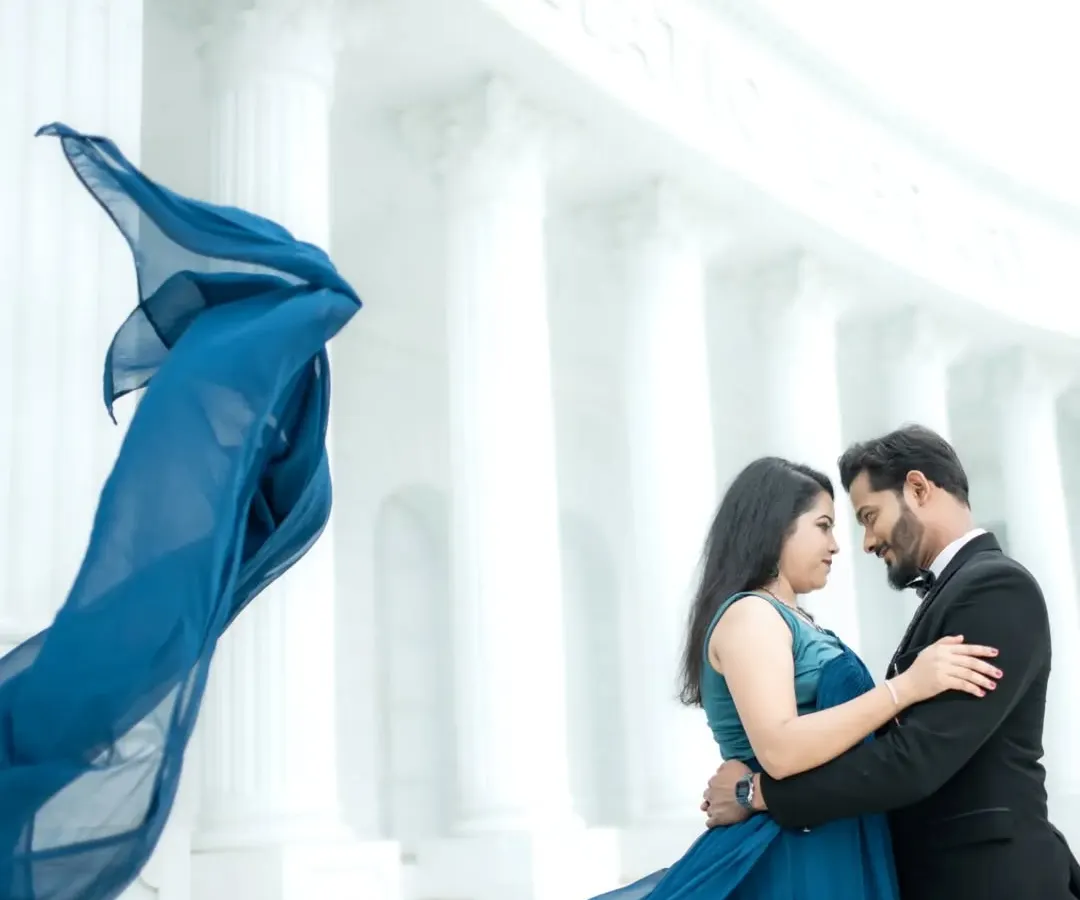 A dramatic, cinematic shot featuring a flowing blue gown against the grand, all-white architecture of a studio set. This style is perfect for couples who dream of a filmi, larger-than-life pre-wedding album.