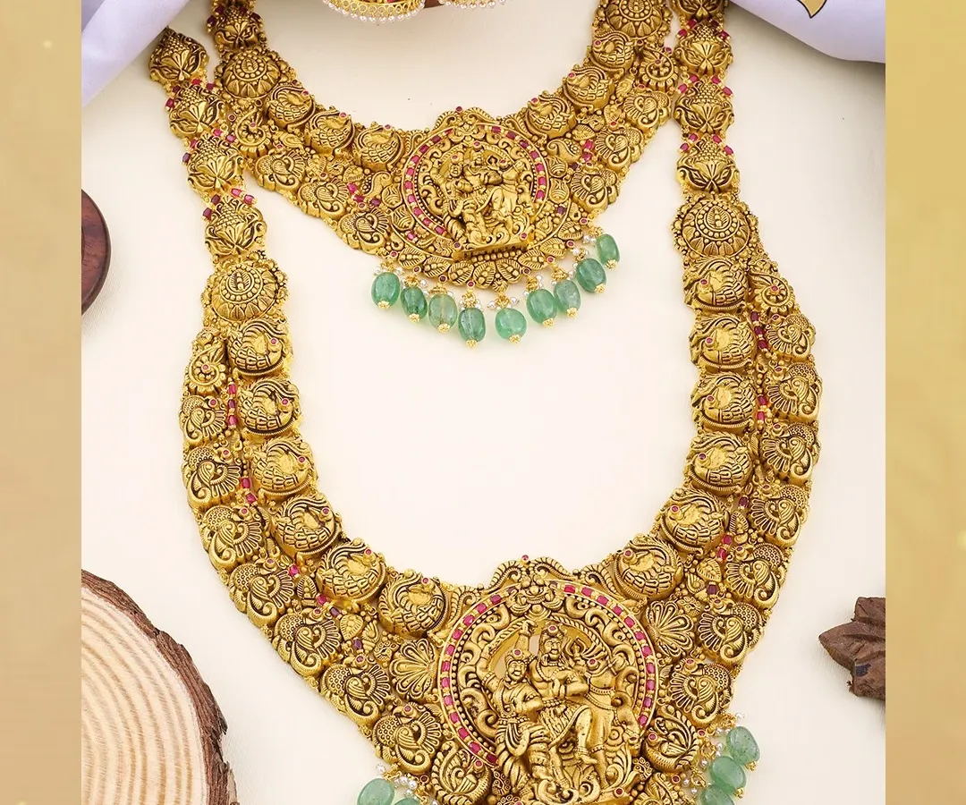 This magnificent double-layered temple jewellery set is a masterpiece of our heritage collection. Crafted in antique-finish 22kt gold with green bead accents, it features detailed carvings of Lord Krishna, making it a highly sought-after piece for discerning bridal customers.