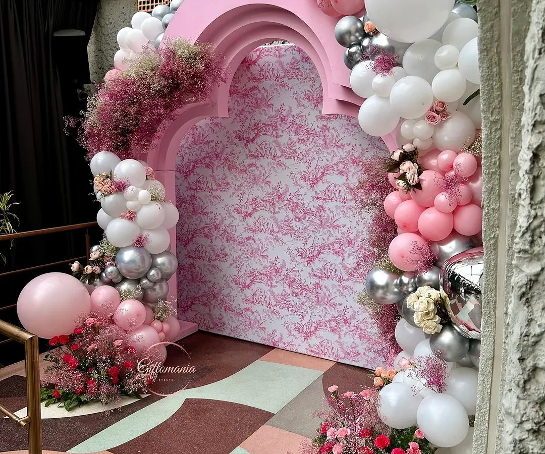 This Dior-inspired 40th birthday party backdrop features a custom-printed panel in a signature pattern, framed by an elegant pink arch. The look is completed with a cascading garland of pink, white, and silver balloons mixed with fresh floral accents.