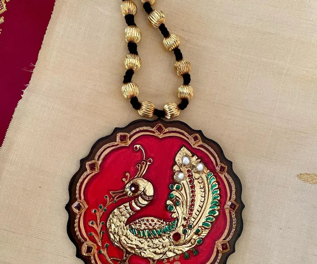 A majestic peacock, brought to life with real Tanjore gold foil against a rich red background. This wearable art piece is sealed with resin to protect its intricate gesso work and brilliant shine, making it a truly royal accessory.