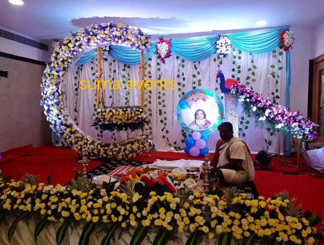 This is a popular naming ceremony stage design featuring a large crescent moon swing made of fresh flowers. The peacock motif and baby's photo add a personal touch to the celebration.