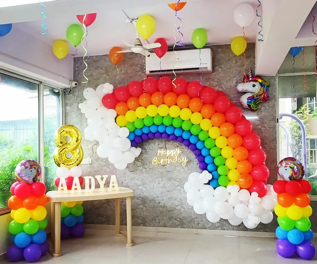 I built this large rainbow balloon arch against a grey wall for an 8th birthday party. The setup includes rainbow-colored balloon pillars, a unicorn foil balloon, and a lit-up 'Happy Birthday' sign to create a bright and cheerful unicorn-themed celebration.