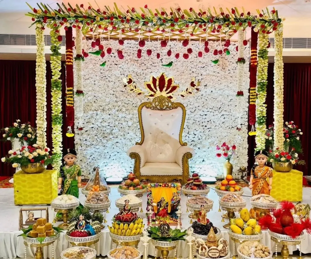 An absolutely divine Seemantham (baby shower) stage, featuring a wall of white flowers, a traditional throne chair, and meticulously arranged offerings. We honour your sacred ceremonies with beauty and respect.