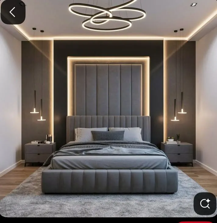 This master bedroom design creates a dramatic and luxurious feel with a floor-to-ceiling upholstered headboard, highlighted by recessed LED backlighting. The layered lighting scheme, including a modern chandelier and hanging bedside pendants, adds depth and sophistication to the contemporary grey palette.