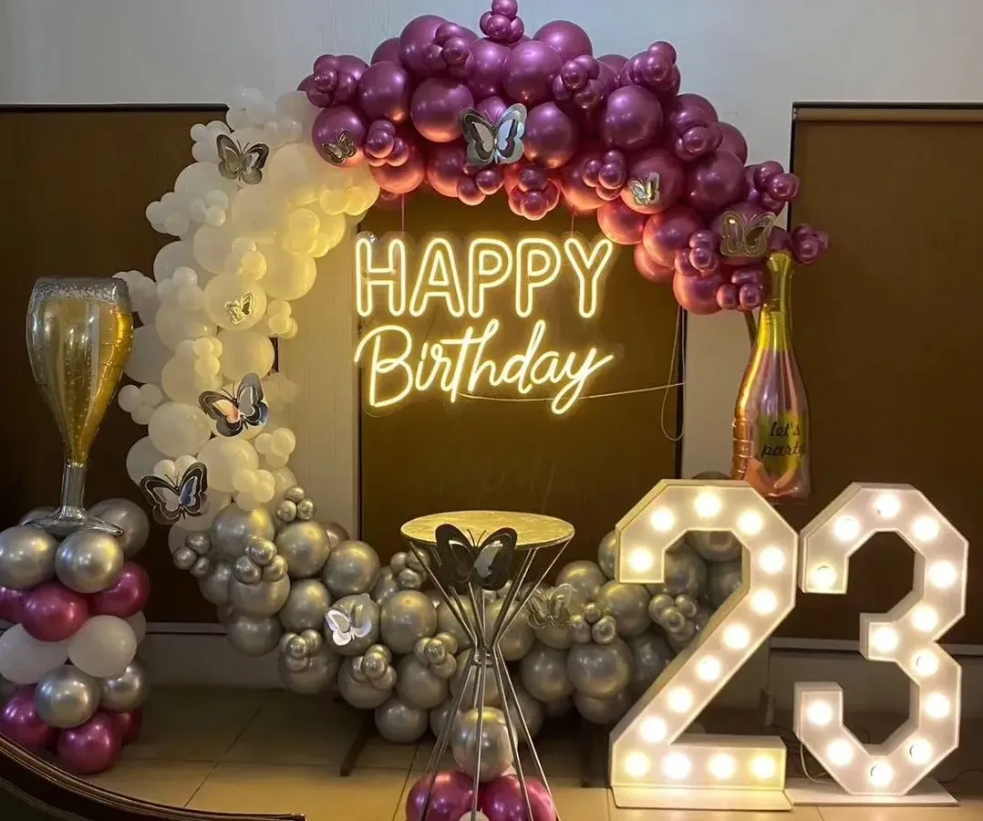 A glamorous 23rd birthday celebration featuring large light-up marquee numbers, a custom "Happy Birthday" neon sign, and a chic balloon arch in magenta, silver, and white with butterfly accents.